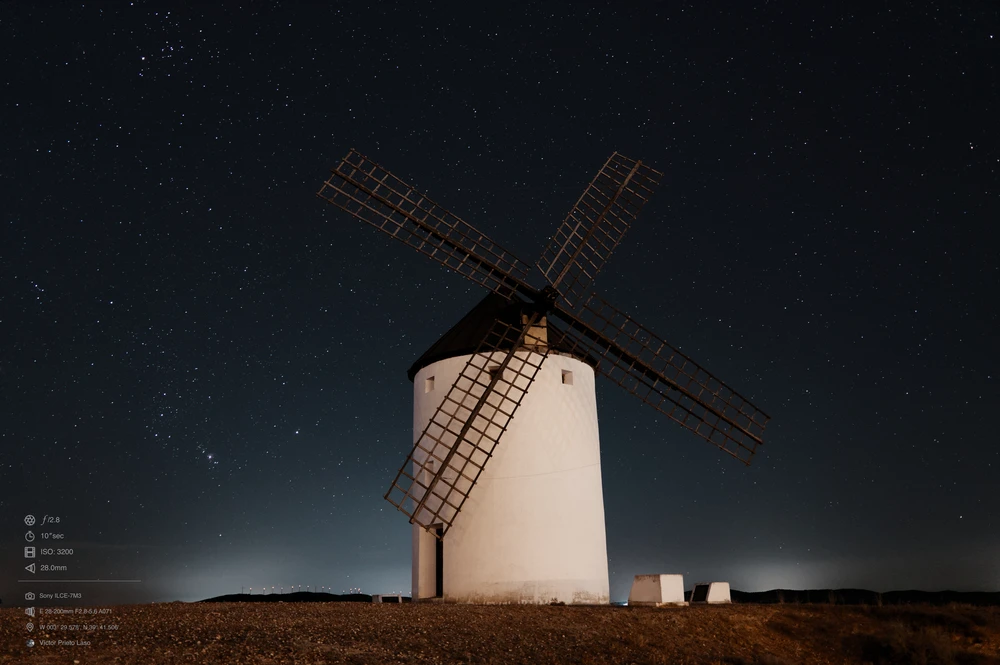 A white windmill with a dark wooden roof and blades sits atop a hill. The long exposure of the photo allows for a clear view of the stars in the night sky. There is some light pollution from nearby villages and the wind farm.