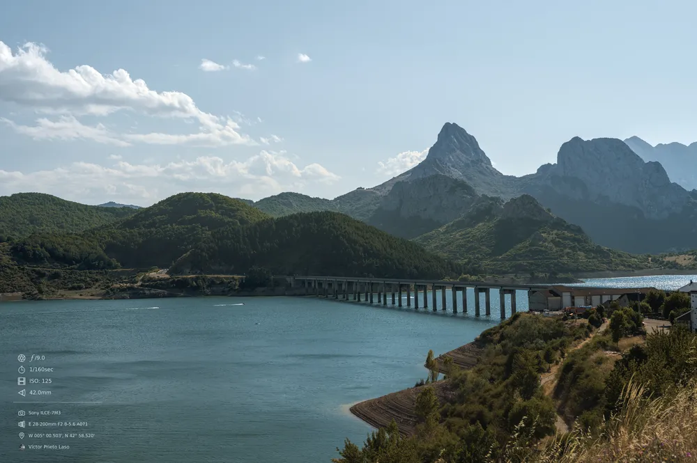 A landscape featuring a long bridge over a water reservoir with green hills and mountains in the background. The sky is clear except for a few clouds. The water is calm, but there are speedboats in the distance. The overall atmosphere is serene and peaceful.