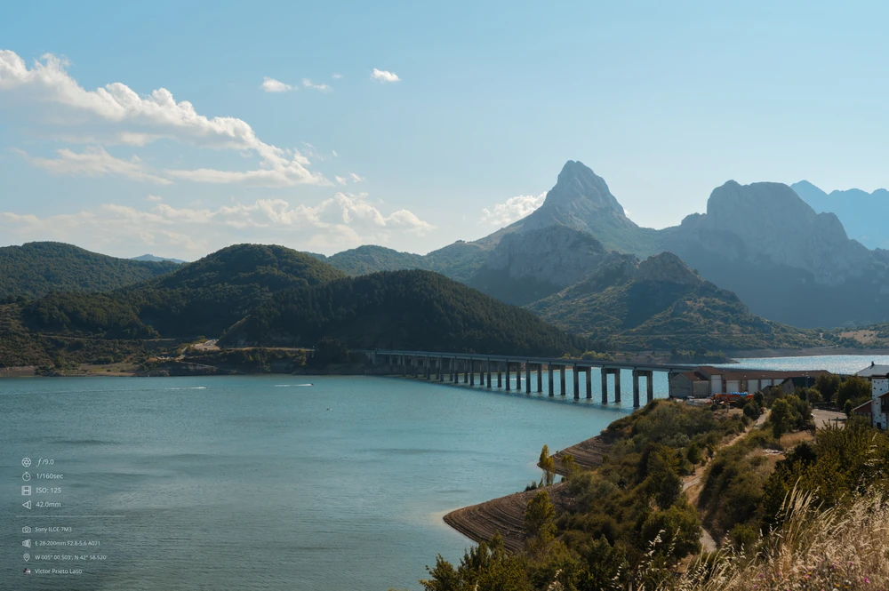 A landscape featuring a long bridge over a water reservoir with green hills and mountains in the background. The sky is clear except for a few clouds. The water is calm, but there are speedboats in the distance. The overall atmosphere is serene and peaceful.